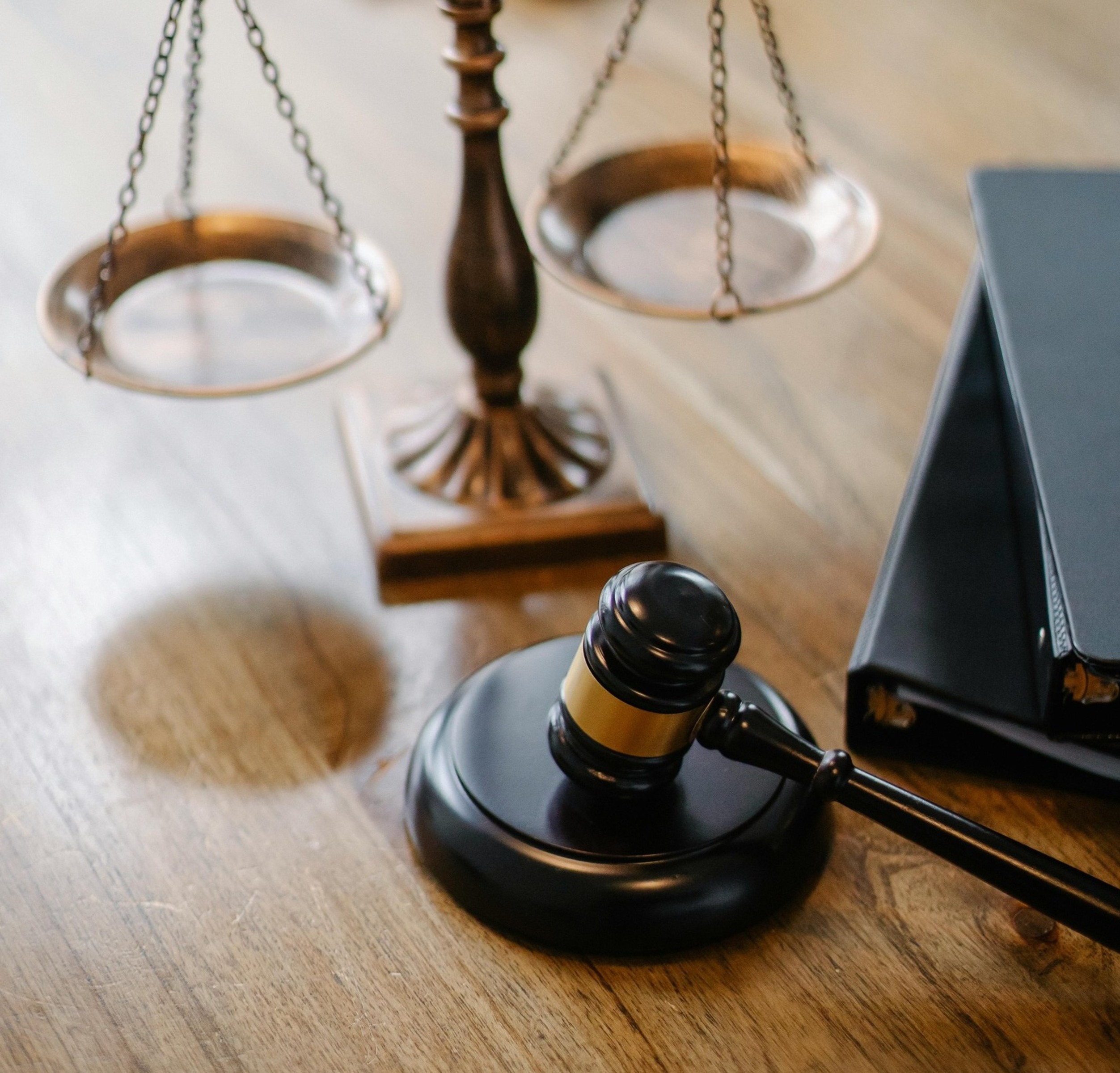 A wooden gavel and bronze balance scale sit on a wooden desk beside legal binders, symbolizing justice and law.