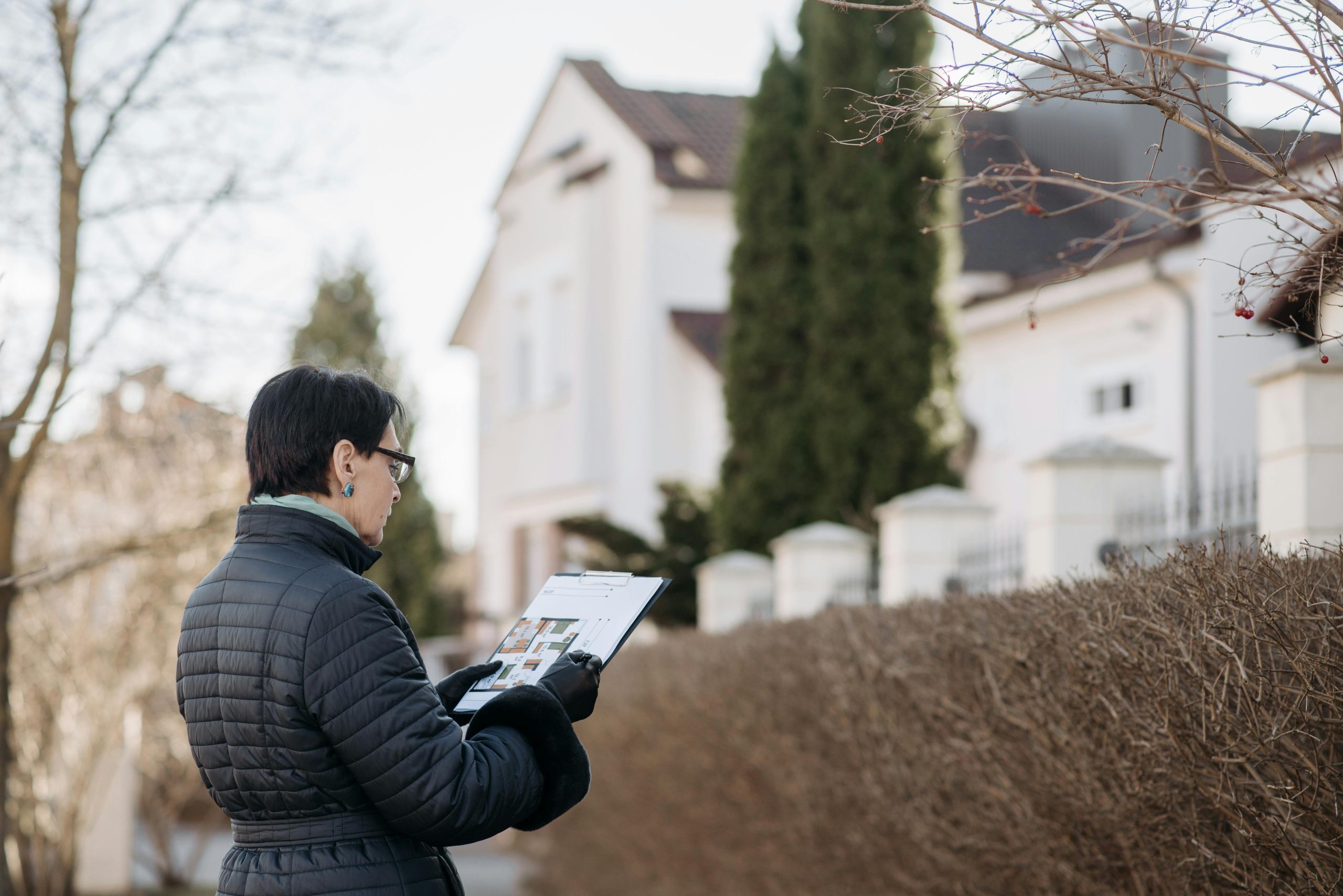 Woman in a black jacket holding a clipboard, inspecting a residential property on a clear day.
